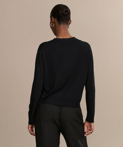 A person with short hair, wearing the black Mira Cardigan and black pants, stands facing away from the camera against a plain beige background.