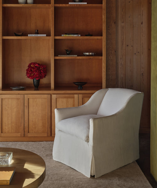 A Miramar Chair Slipcover in white pearl linen is placed on a beige rug near wooden built-in shelves with minimal decor and red flowers. A round wooden table appears partially in the foreground.
