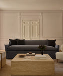 A minimalist living room with a Miramar Sofa in dark gray, two black cushions, a light wood coffee table with books and decor, a plant, cream-hued walls with molding, and a closed white door in the background.