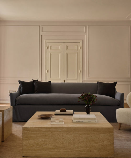 A minimalist living room with a Miramar Sofa in dark gray, two black cushions, a light wood coffee table with books and decor, a plant, cream-hued walls with molding, and a closed white door in the background.
