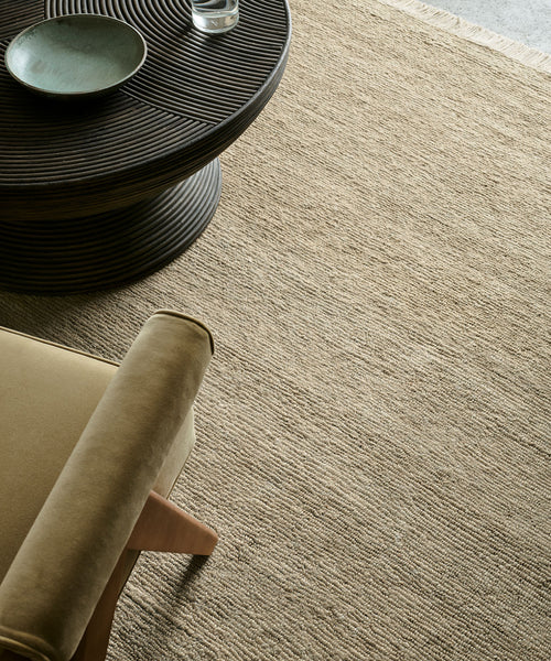 A round dark wooden coffee table with a bowl and glass sits next to a beige upholstered armchair on the Mojave Jute Rug, a textured light brown rug handmade in India.