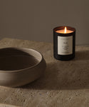 The Musk Glass Candle, hand-poured in Los Angeles with coconut soy wax, rests on a textured stone surface next to a neutral ceramic bowl, creating a calm and minimalist scene.