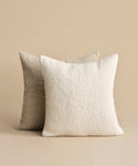 The Nell Boucle Pillow, featuring a soft ivory boucle texture, is displayed in front of a light brown textured pillow against a beige background.