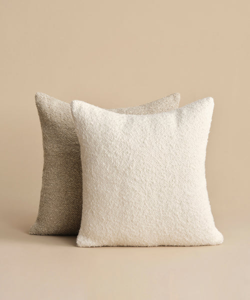 The Nell Boucle Pillow, featuring a soft ivory boucle texture, is displayed in front of a light brown textured pillow against a beige background.