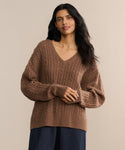 A person with long, dark hair wears the Nell V-Neck Sweater in a relaxed fit and brown hue, paired with dark pants, standing against a plain beige background with a calm expression.