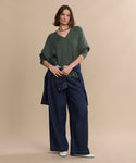 A woman stands against a beige background wearing the Nell V-Neck Sweater in green, wide-leg blue jeans, a blue jacket tied around her waist, white shoes, and a choker necklace.