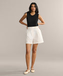 A woman with long, dark hair poses against a plain background, hands on hips, wearing a sleeveless black top, Newport Short in Italian cotton poplin, and cream flat shoes.