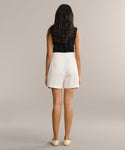 A woman with long dark hair, in a sleeveless black top and Newport Short, stands facing away against a beige background—showcasing effortless summer shorts style.