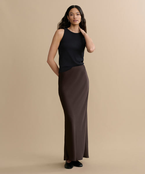 A woman stands against a beige background wearing a sleeveless black top, black shoes, and the Nina Slip Skirt in brown. She has long wavy hair, with one hand at her side and the other touching her hair.