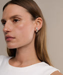 A woman with straight brown hair wears chunky Noodle Earrings and a sleeveless white top, gazing slightly to the left against a neutral beige background.