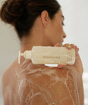 A woman with wet hair lathers Awaken Body Wash on her shoulder and back. The white pump bottle, which contains Rice Bran Extract, is visible as she looks away from the camera.