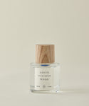A clear glass bottle of Santo Wood Eau De Parfum with a wooden cap rests on a light beige surface. Its label shows 50 ml (1.7 fl oz). The clean, non-toxic fragrance stands out against the plain, light-colored background.