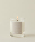 The Violet Santal Candle, in a clear glass jar with a beige label, fills the air with fresh violet, crisp amber, and California woods for coastal nostalgia against a light background.