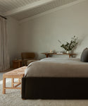 Minimalist bedroom with a dark bed, neutral bedding, the Montecito Bench in white oak, small table with greenery in a vase, chair, and beige curtains. Soft natural light and pale walls create a calm atmosphere.