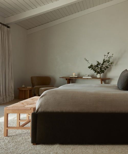 Minimalist bedroom with a dark bed, neutral bedding, the Montecito Bench in white oak, small table with greenery in a vase, chair, and beige curtains. Soft natural light and pale walls create a calm atmosphere.
