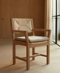 A Montecito Dining Arm Chair with a Belgian linen cushion and wooden frame sits on a tiled floor in a minimalist room featuring wood-paneled walls and large windows draped with sheer curtains.