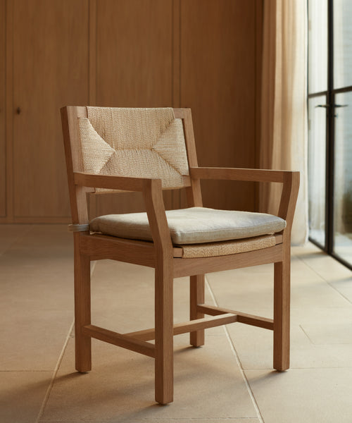 A Montecito Dining Arm Chair with a Belgian linen cushion and wooden frame sits on a tiled floor in a minimalist room featuring wood-paneled walls and large windows draped with sheer curtains.