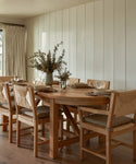 A wooden dining table set for six features the Montecito Dining Arm Chair, each with a Belgian linen cushion. Neutral dinnerware, glasses, and napkins enhance the vase centerpiece in a space with light wood walls and curtains.