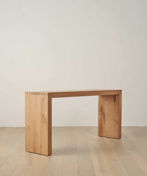 The Malibu Console Table features a minimalist design with a rectangular White Oak top and two thick vertical legs with subtle box joint details, shown on a light wood floor against a plain white wall.