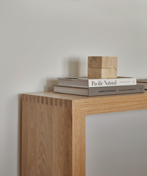 A close-up of the Malibu Console Table in White Oak features box joint detailing and is styled with two books, a magazine, and a small stone ornament, set against a plain white wall.