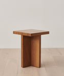 The Outdoor Marin Square Side Table features a small, modern teak design with a square top and three intersecting vertical panels as its base, styled on a light wood floor against a plain white wall.