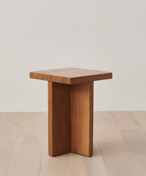 The Outdoor Marin Square Side Table features a small, modern teak design with a square top and three intersecting vertical panels as its base, styled on a light wood floor against a plain white wall.