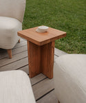 The Outdoor Marin Square Side Table with a modern, minimalist design sits on a wooden deck among beige cushioned chairs, grass in the background. Four round stone coasters are arranged on the tables square top.