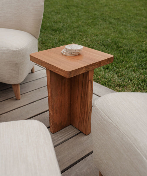 The Outdoor Marin Square Side Table with a modern, minimalist design sits on a wooden deck among beige cushioned chairs, grass in the background. Four round stone coasters are arranged on the tables square top.