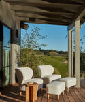 A covered wooden patio featuring two white chairs with matching ottomans, the Outdoor Marin Square Side Table, and a scenic view of green hills and blue sky in the background.