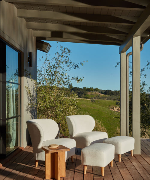 A covered wooden patio featuring two white chairs with matching ottomans, the Outdoor Marin Square Side Table, and a scenic view of green hills and blue sky in the background.