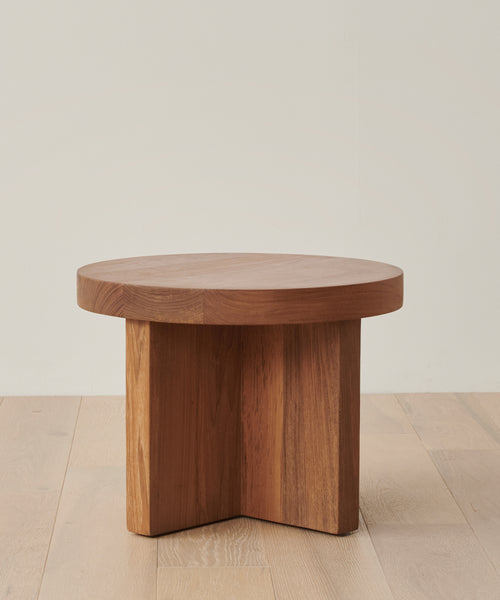 A round wooden coffee table with a thick top and intersecting base sits on a light wood floor against an off-white wall—perfectly protected year-round by the Outdoor Marin Low Side Table Cover.