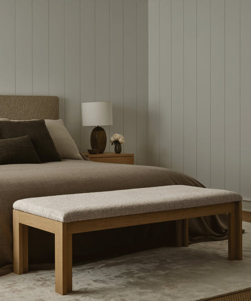 A modern bedroom features a beige upholstered bed with brown bedding, the Otto Bench in wool boucle at the foot, a bedside table with lamp, and neutral-toned walls and rug.