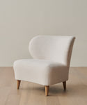 The Outdoor Brentwood Chair, a modern armless accent chair with light beige upholstery and wooden legs, is handmade in Los Angeles and displayed on a light wood floor against an off-white wall.