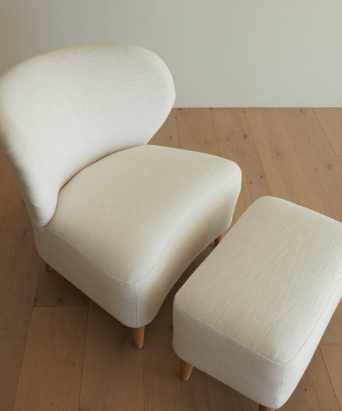 The Outdoor Brentwood Chair, cream-colored and upholstered with a curved backrest, is handmade in Los Angeles and paired with a matching rectangular ottoman. Both have wooden legs and are set on a light wooden floor.