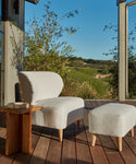 An Outdoor Brentwood Chair in performance ivory with a matching ottoman is placed on a wooden patio next to a small wooden side table, overlooking green hills and trees under a clear blue sky.