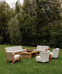 A modern outdoor seating area on grass features handmade wooden furniture from Los Angeles—a sofa, two armchairs, and Outdoor Brentwood Chairs with performance ivory cushions—arranged around two wooden coffee tables amid trees.