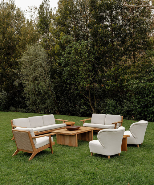 A modern outdoor seating area on grass features handmade wooden furniture from Los Angeles—a sofa, two armchairs, and Outdoor Brentwood Chairs with performance ivory cushions—arranged around two wooden coffee tables amid trees.