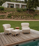 Two cream-colored Outdoor Brentwood Chairs with performance textured linen and solid teak legs, a matching ottoman, and a wooden side table are arranged on the deck by the pool, with views of a grassy lawn and modern house.