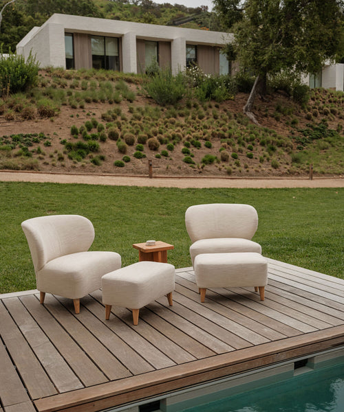 Two cream-colored Outdoor Brentwood Chairs with performance textured linen and solid teak legs, a matching ottoman, and a wooden side table are arranged on the deck by the pool, with views of a grassy lawn and modern house.