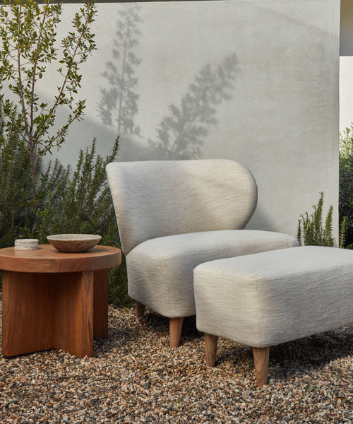 A modern outdoor space showcases the Outdoor Brentwood Chair with solid teak legs and light gray cushions, paired with a matching ottoman on gravel beside a wooden table, surrounded by lush plants and a light-colored wall.