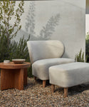 A modern outdoor space includes a light gray cushioned chair with solid teak legs and the Outdoor Brentwood Ottoman on gravel, beside a wooden side table with ceramic bowls, all set amid greenery against a pale wall.