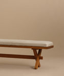 The Outdoor Garden Dining Bench, crafted from solid teak, features angled crisscross legs and includes a light beige Tempotest fabric cushion. It is shown on a beige floor with a matching background.