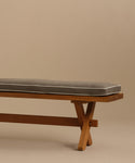 The Outdoor Garden Dining Bench features crossed solid teak legs and a gray Tempotest fabric cushion, shown against a plain beige background.