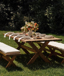 The Outdoor Garden Dining Table is arranged with white dishes, glasses, and napkins. A floral centerpiece adds charm as the table sits on green grass with trees in the background. Matching benches complete the garden dining setup.