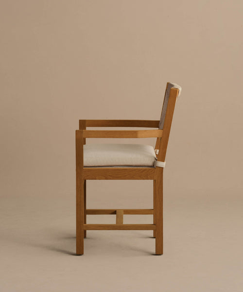 The Outdoor Montecito Dining Arm Chair, crafted from solid teak with a light beige cushioned seat and backrest, is shown in a side view against a plain beige background, highlighting its simple modern design and clean lines.