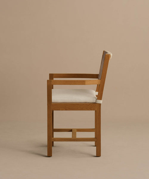 The Outdoor Montecito Dining Arm Chair is displayed in profile against a plain light brown background, featuring clean, straight lines and a modern design with a wooden frame and light beige Tempotest fabric cushion and backrest.