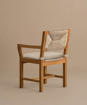 The Outdoor Montecito Dining Arm Chair features solid teak, a light beige Tempotest fabric cushion, and a woven backrest. Its simple, modern design highlights the natural wood grain and clean lines against a taupe background.