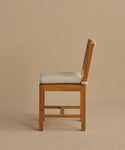 The Outdoor Montecito Dining Chair features a teak frame and light beige cushioned seat and backrest. Shown in profile against a plain, light brown background, its modern design offers style and durability for outdoor dining spaces.