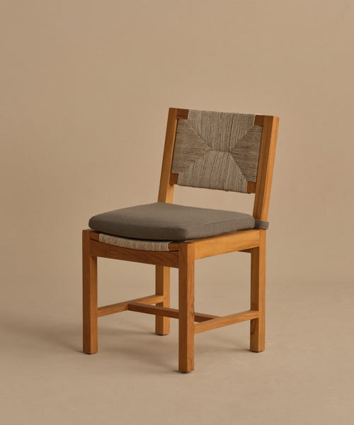 The Outdoor Montecito Dining Chair features a modern design with a cushioned gray seat, woven ECOLENE backrest, and solid teak frame. Set against a plain beige background, its clean lines and simple style create an elegant outdoor seating option.