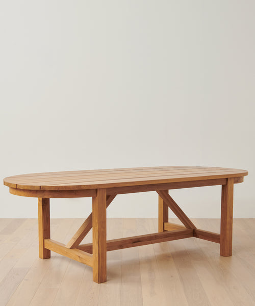 The Outdoor Montecito Oval Dining Table, featuring a slatted teak top and sturdy cross-braced legs, stands on a light wood floor against a white wall—an elegant centerpiece for indoor or outdoor dining.